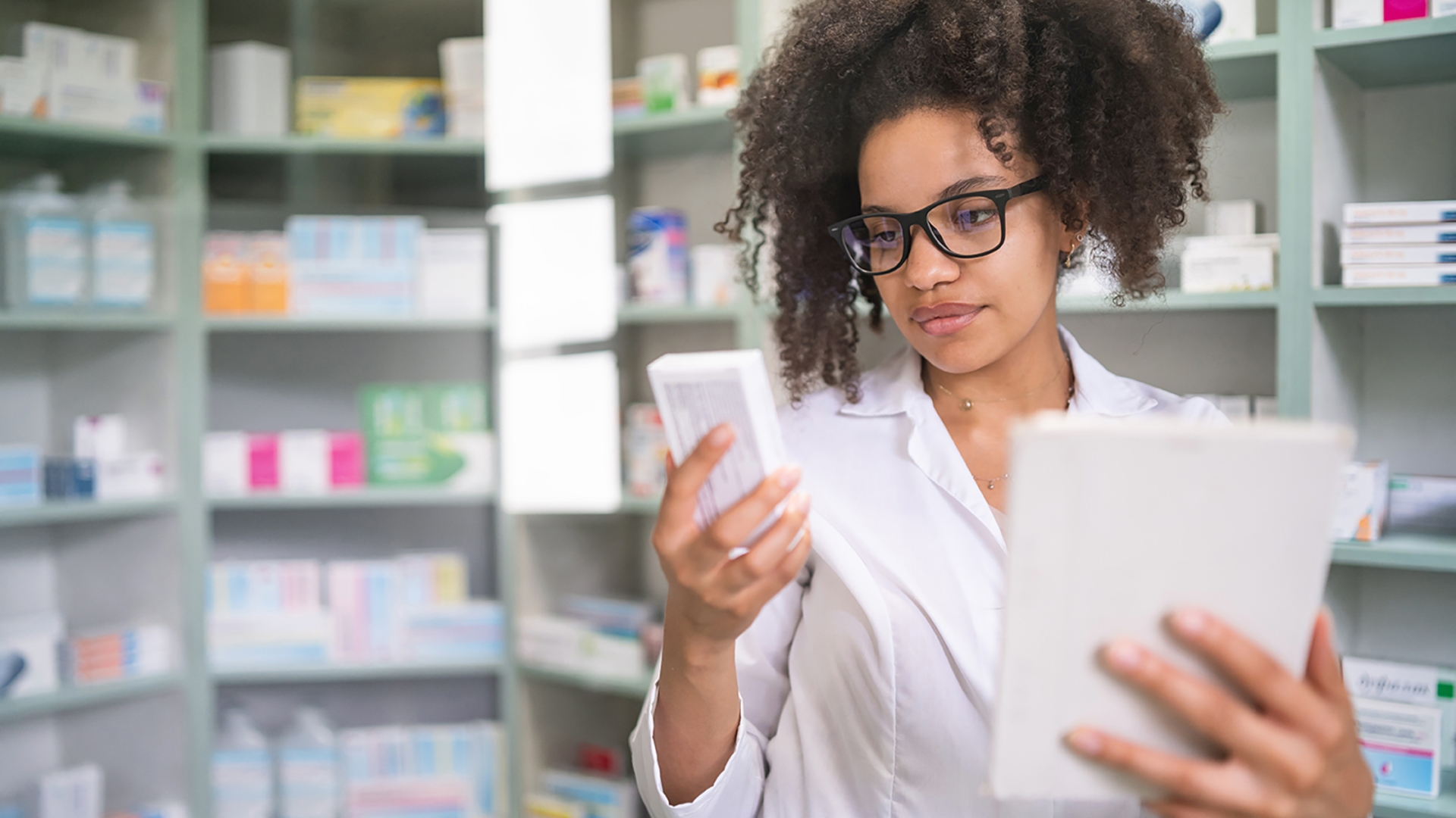 Person looking at a box of medication with a tablet in their hands standing in front of a shelf of medication.
