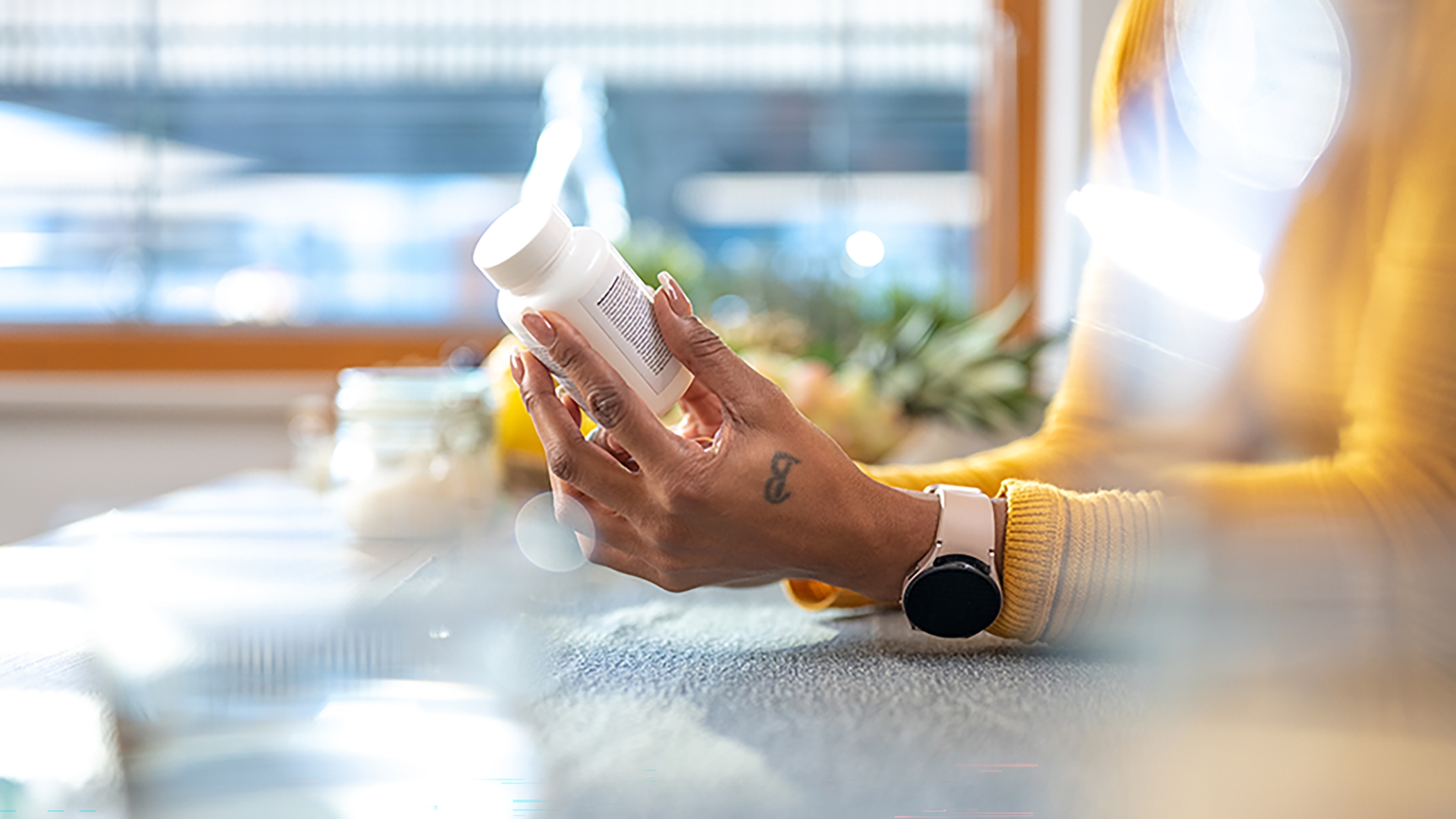 Person reading the label on a white pill bottle, wearing a yellow sweater and smartwatch, seated at a table with blurred background.